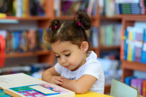 Little Girl Indoors In Front Of Books. Cute Young Toddler Sitting On A Chair Near Table and Reading Book. Child reads in a bookstore, surrounded by colorful books. Library, Shop, Shelving In Home. The Library is one of many free parenting resources in saint louis.